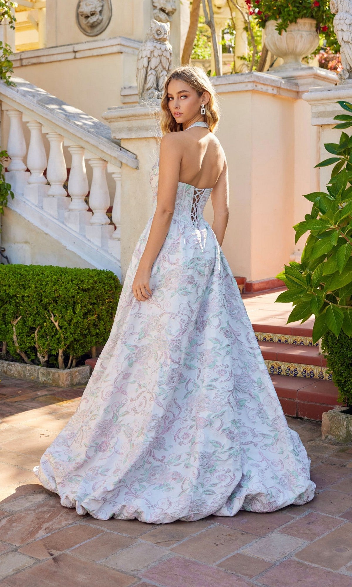 A woman in the Juliet JT2742A long prom dress stands outdoors on a tiled patio near white stairs and greenery, looking over her shoulder.